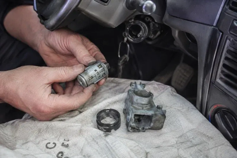 Hands holding a removed ignition cylinder beside a car steering column, with the housing and small parts on a cloth. — Ignition Lock Repair, Peterborough