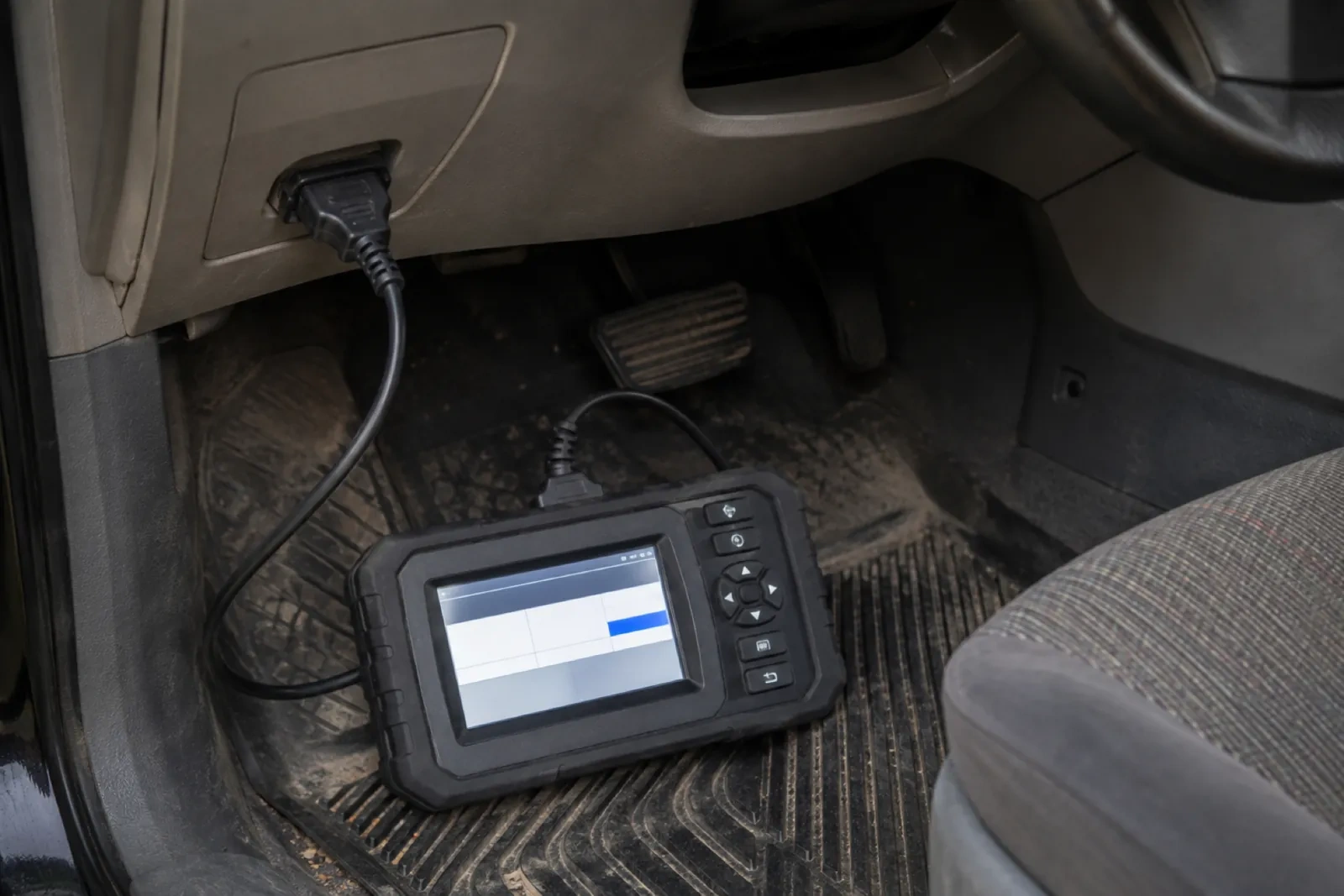 Handheld diagnostic unit with a screen and buttons lying on a dirty car floor mat, cabled to a port under the dash.